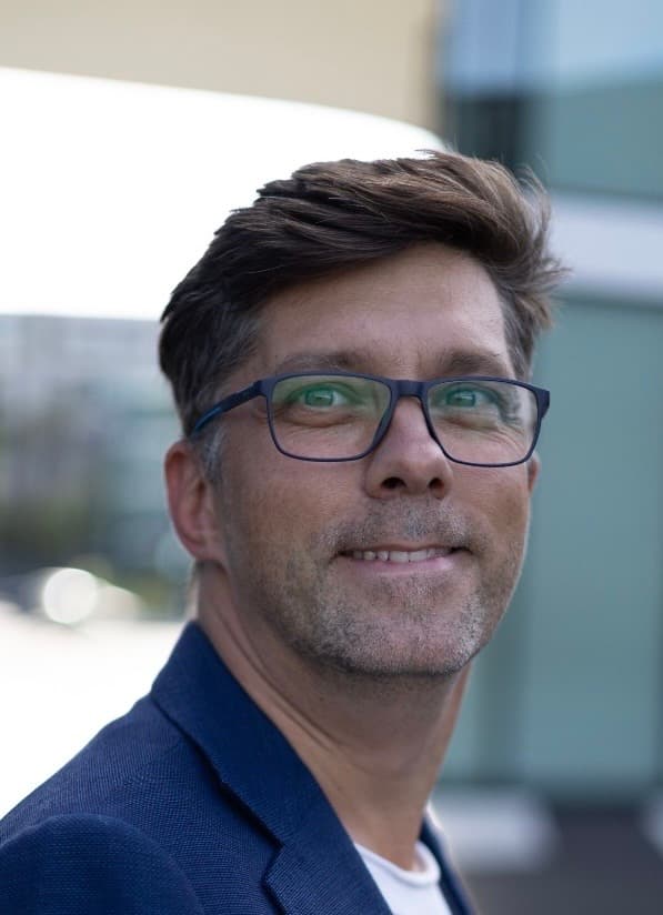 Ein lächelnder Mann mit braunem Haar und Brille trägt ein blaues Sakko im Freien.