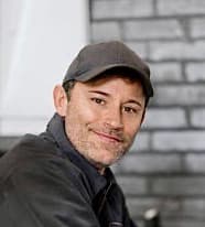 Lächelnder Mann mit Bartstoppeln und grauer Kappe vor einer unscharfen grauen Ziegelwand im Hintergrund.