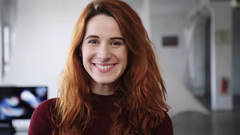 Lächelnde Frau mit langen roten Haaren und dunkelrotem Oberteil blickt direkt in die Kamera.