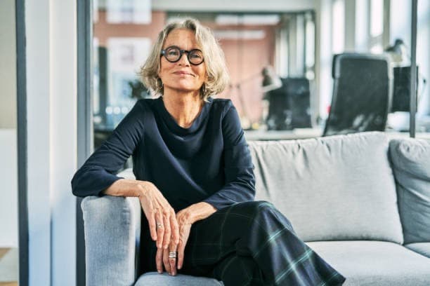 Reife Frau mit Brille und kurzen grauen Haaren sitzt lächelnd auf einem Sofa im Büro.
